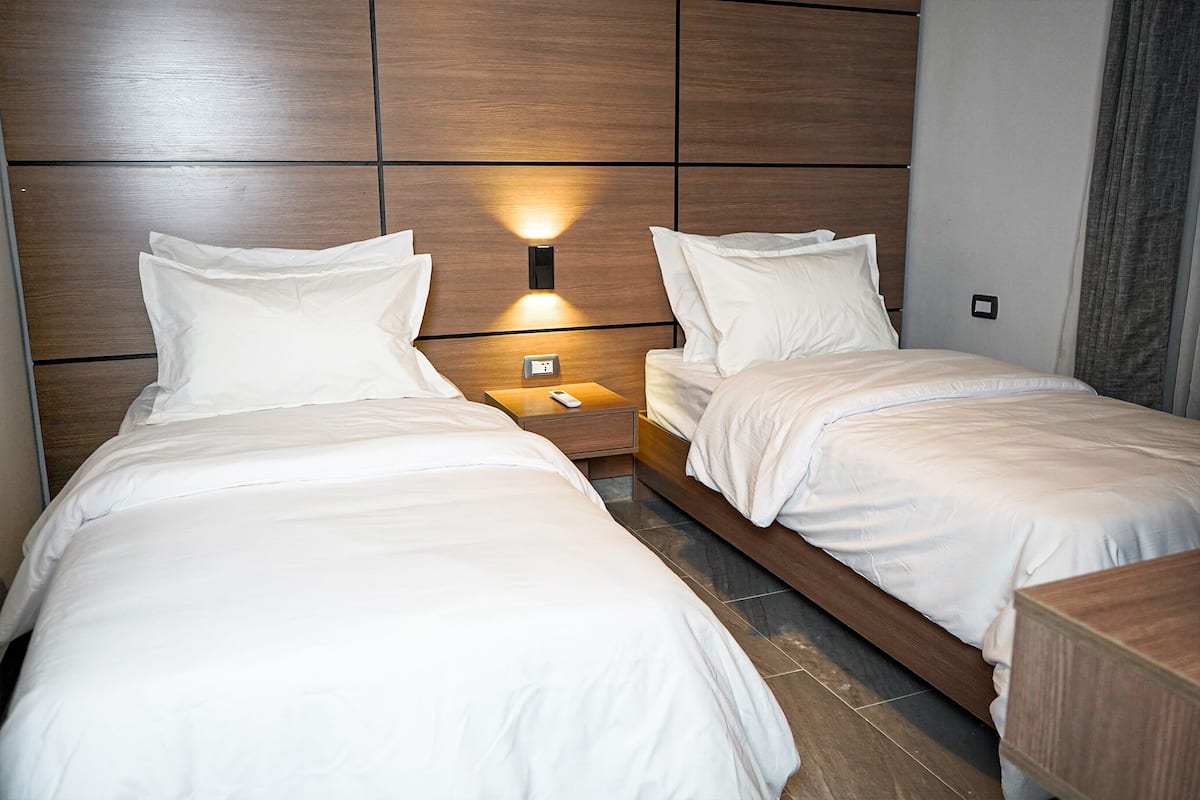 Habitación de hotel con dos camas individuales, cabecero de madera, mesitas de noche y una lámpara fijada a la pared.