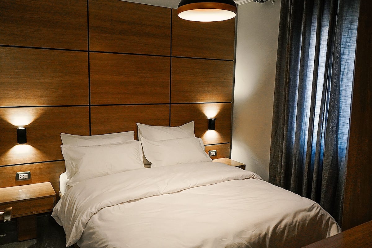 Una cama bien hecha con ropa de cama blanca en una habitación con paneles de madera y dos luces fijadas a la pared.