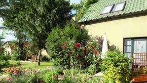 Ein Garten mit verschiedenen Pflanzen und Blumen, einer Holzschaukel und einem Haus mit grüner Dachbegrünung und einem Fenster.