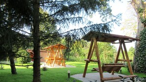 Ein hölzernes Schaukelgerüst und ein Holzspielhaus in einer grünen, von Bäumen gesäumten Außenanlage.