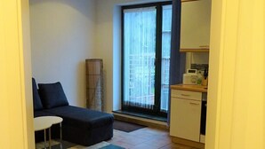 Ein kompakter Wohnbereich mit einer Couch, einem kleinen Tisch, einer Küchenzeile mit Schränken und einem großen Fenster mit Jalousien.