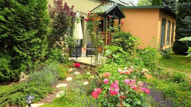 Ein Garten mit verschiedenen Pflanzen und Blumen, eine Terrasse mit Gartenmöbeln und ein Haus mit Satteldach.