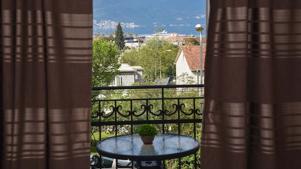 Un balcon avec une table ronde et un pot de fleurs, d’où l’on peut admirer un paysage urbain avec des bâtiments et une montagne à l’horizon.