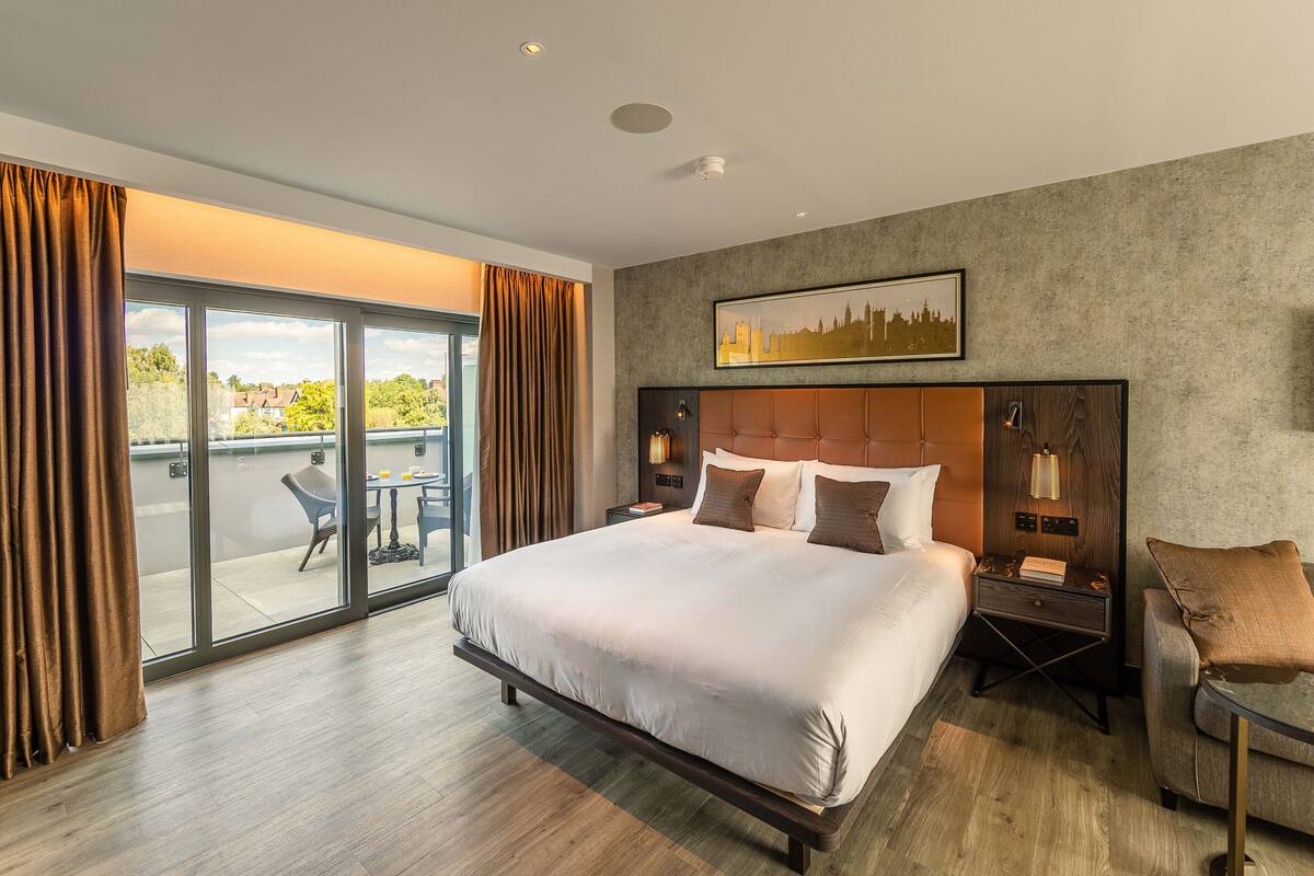 Quarto de hotel moderno com uma cama grande, uma mesa de cabeceira, uma cadeira e uma varanda com mesa e cadeiras.