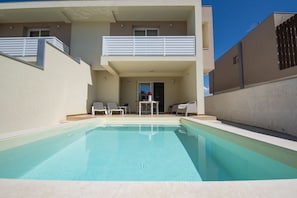 A modern two-story building with a swimming pool, outdoor seating, and a balcony.