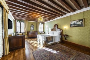 Una habitación con techo de madera, una cama con cabecera, un candelabro, un ventanal grande y un cuadro en la pared.