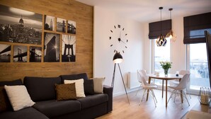 Ett modernt vardagsrum med en svart soffa, en väggpanel i trä, en väggmålning som föreställer en stadsbild, en svart klocka, en matplats med bord och stolar samt en golvlampa.