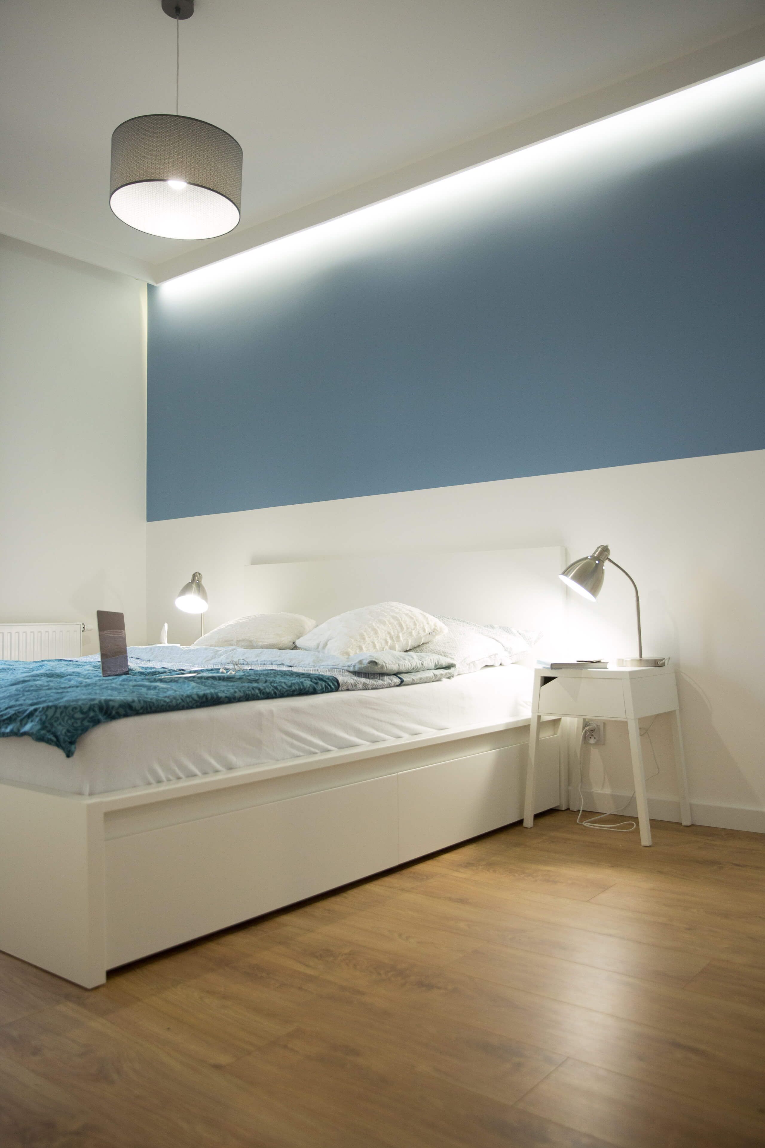 Ein modernes Schlafzimmer mit einem weißen Bett, einer blauen Akzentwand, einer Pendelleuchte und einem Nachttisch mit einer Lampe.