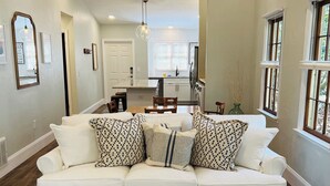 A spacious living area with a white sofa, a matching ottoman, and a patterned rug. A kitchen is visible in the background, and there are windows with blinds.