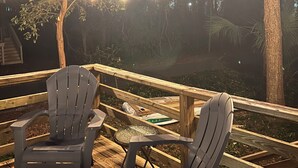 A wooden deck with two Adirondack chairs, string lights, and a view of trees at dusk.