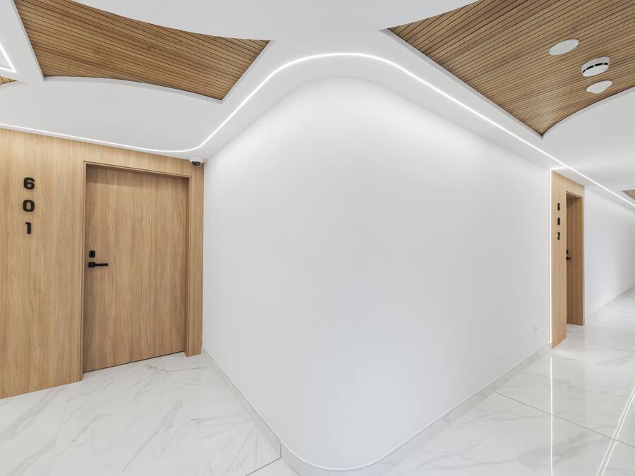 Un pasillo moderno con puertas de madera marcadas con el número “601”, techo curvo y suelo blanco brillante.