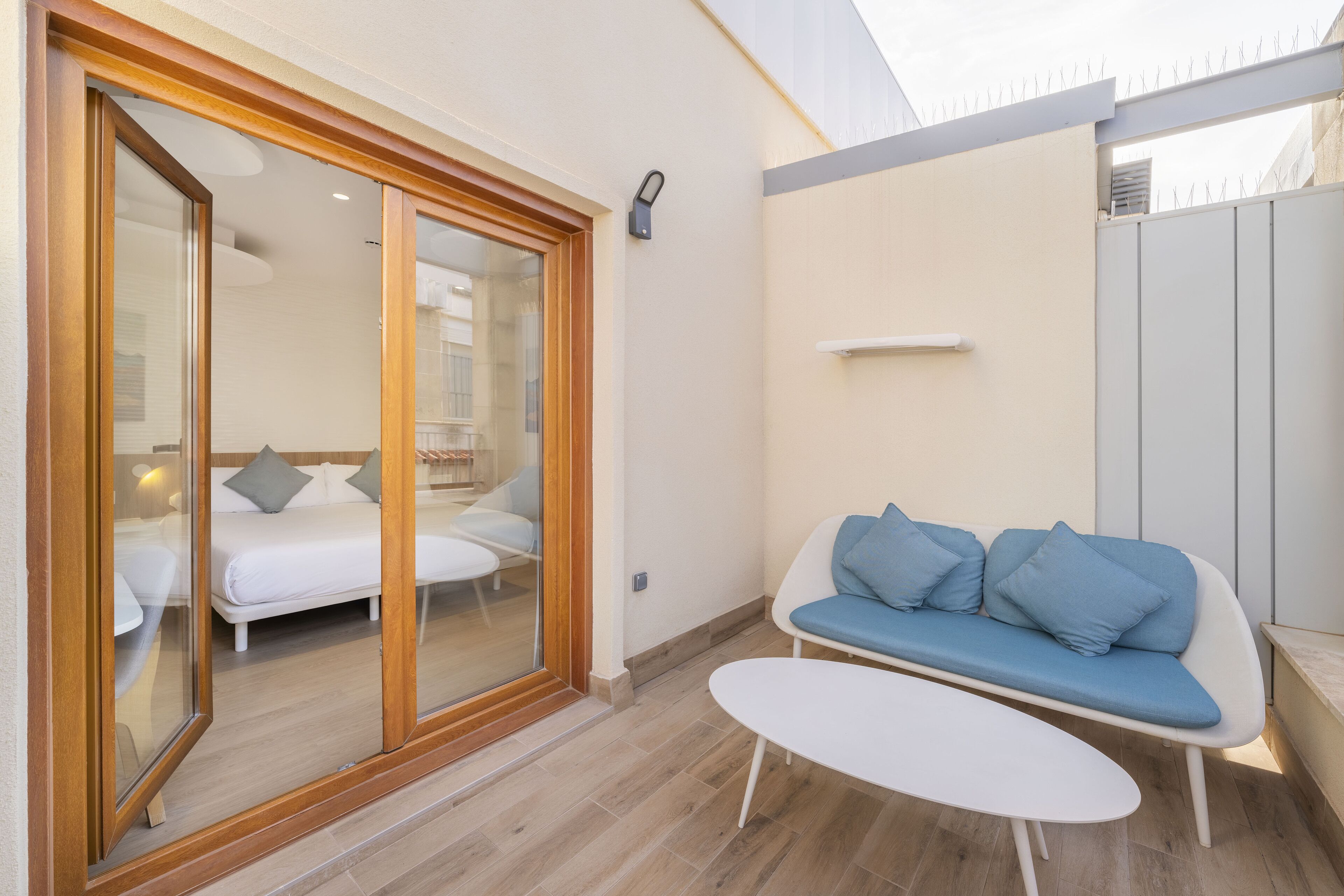 Una habitación moderna con una puerta corredera de vidrio que da a un dormitorio, un sofá blanco con cojines azules y una pequeña mesa blanca.