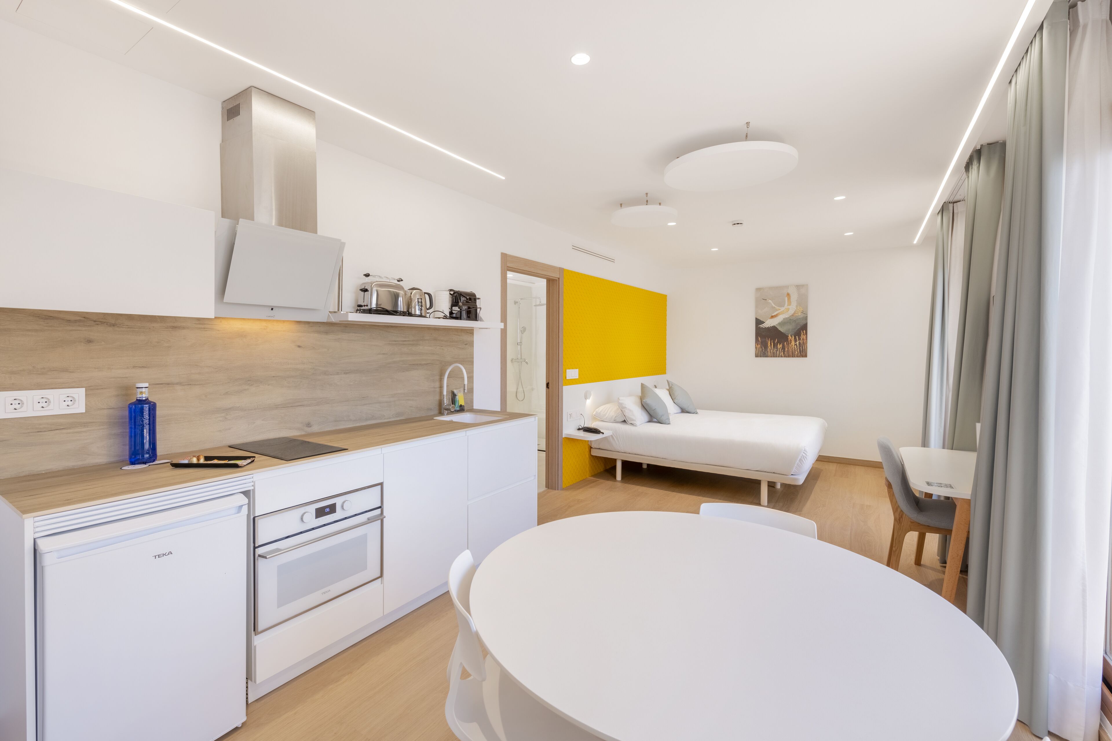 A modern kitchen with a refrigerator, oven, and a sink. There is a dining area with a table and chairs. The room has a bed with a white headboard and a yellow accent wall.