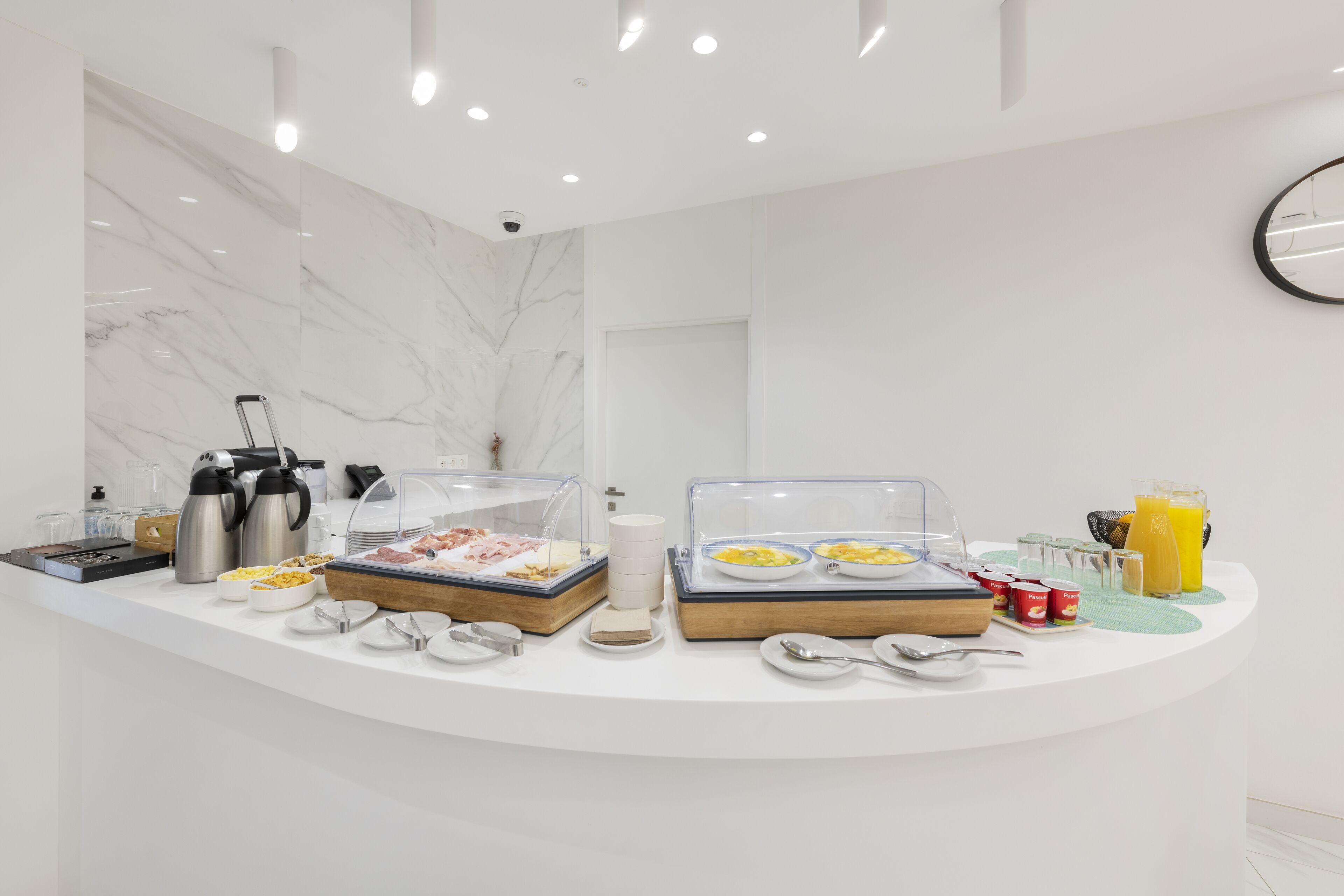 A breakfast buffet with various food items and beverages, a coffee maker, and a clock on the wall.