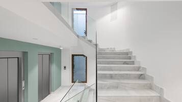 Un interior moderno con una escalera de mármol, barandillas de vidrio y vistas al cielo a través de una ventana.