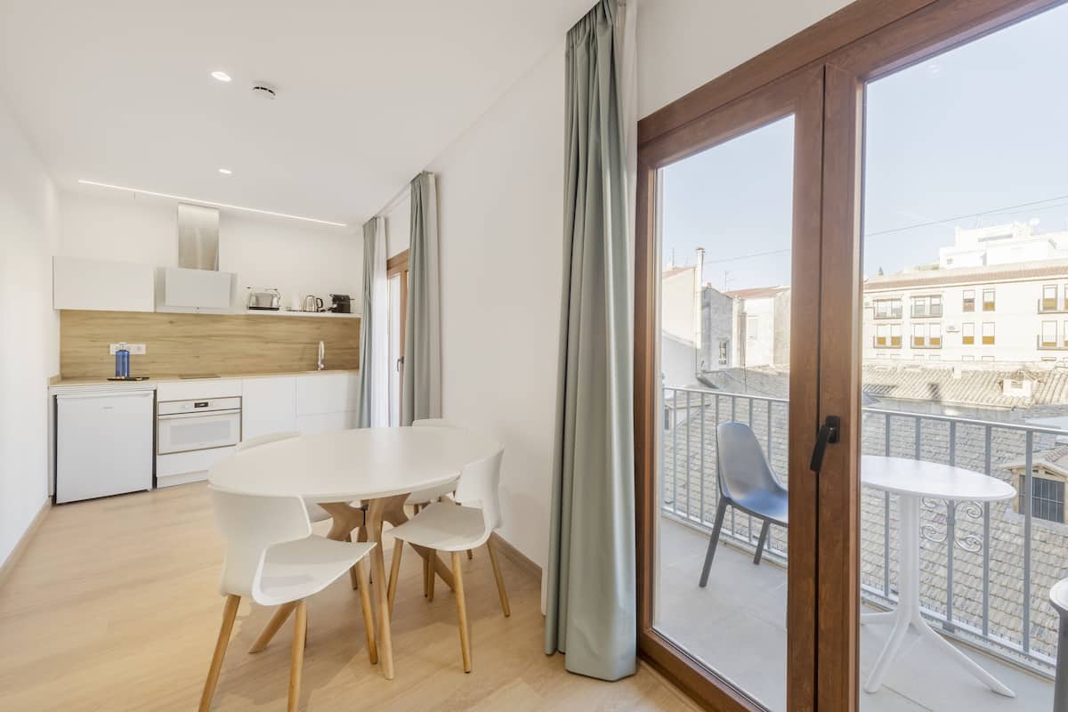 Eine moderne Küche mit weißen Schränken, einem eingebauten Ofen und einem kleinen Kühlschrank. Es gibt einen Essbereich mit einem weißen Tisch und Stühlen. Durch eine Glastür ist ein Balkon mit Geländer und Blick auf Gebäude zu sehen.