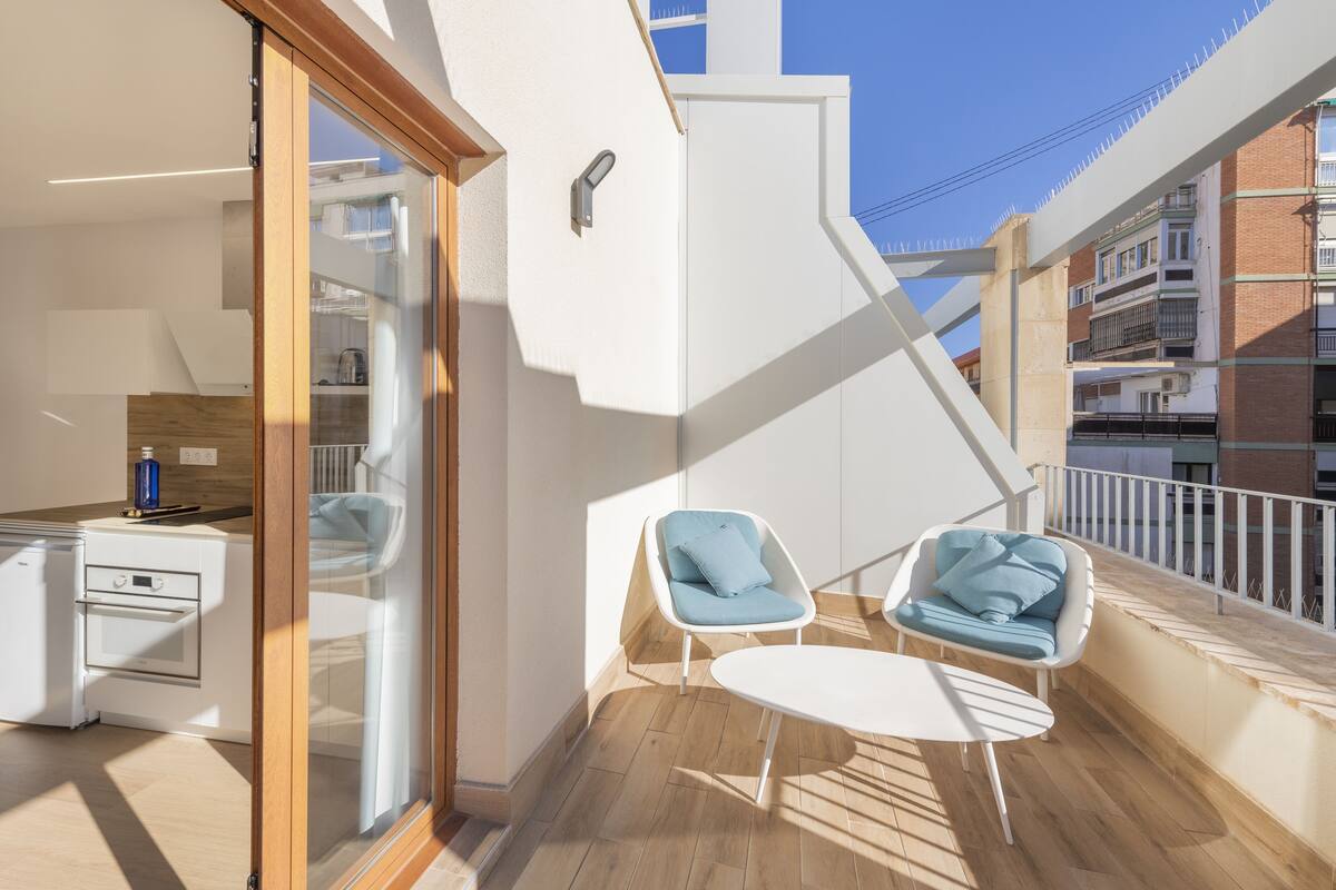 Ein Balkon mit einem weißen Tisch und zwei blauen Stühlen, eine Küchenzeile mit einem weißen Backofen und Blick auf benachbarte Gebäude.