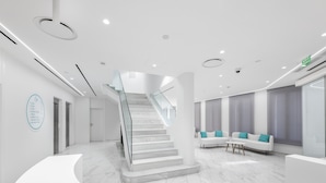 Un área de recepción moderna con un mostrador curvo, una escalera de cristal y un espacio de descanso con sofás blancos y cojines color turquesa.