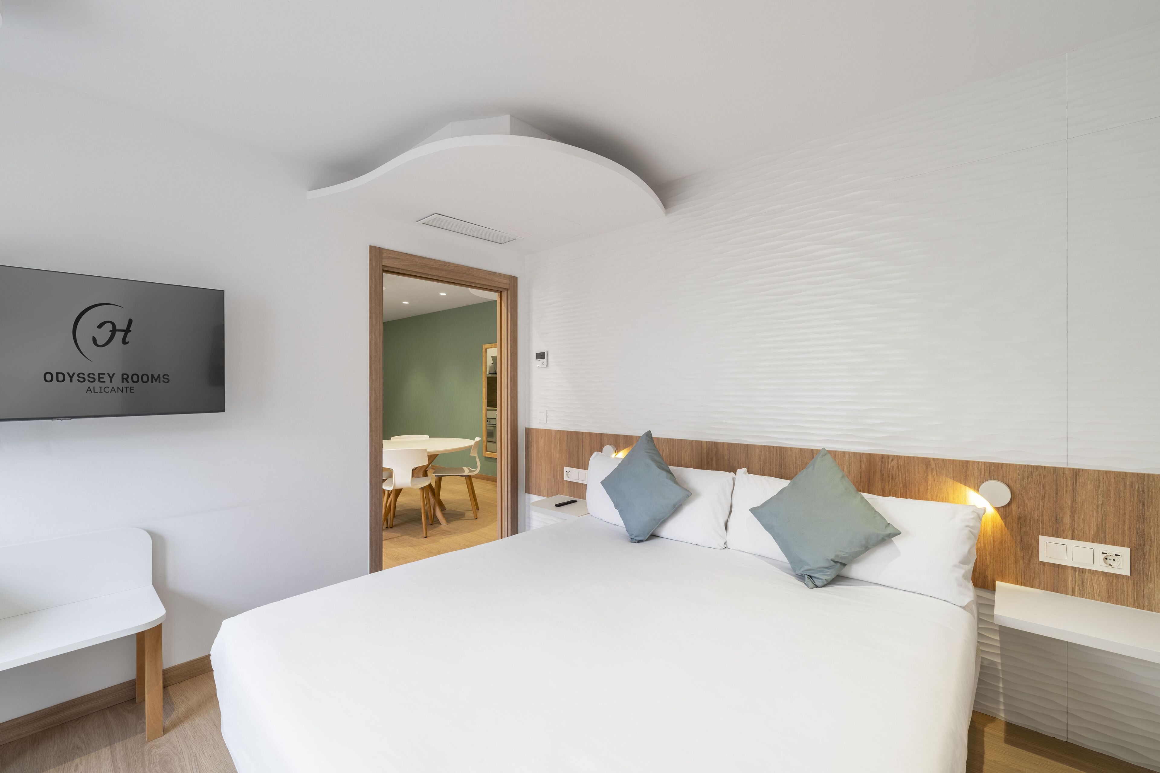 A modern hotel room with a large bed, bedside tables, a flat-screen TV, and a view of the dining area.