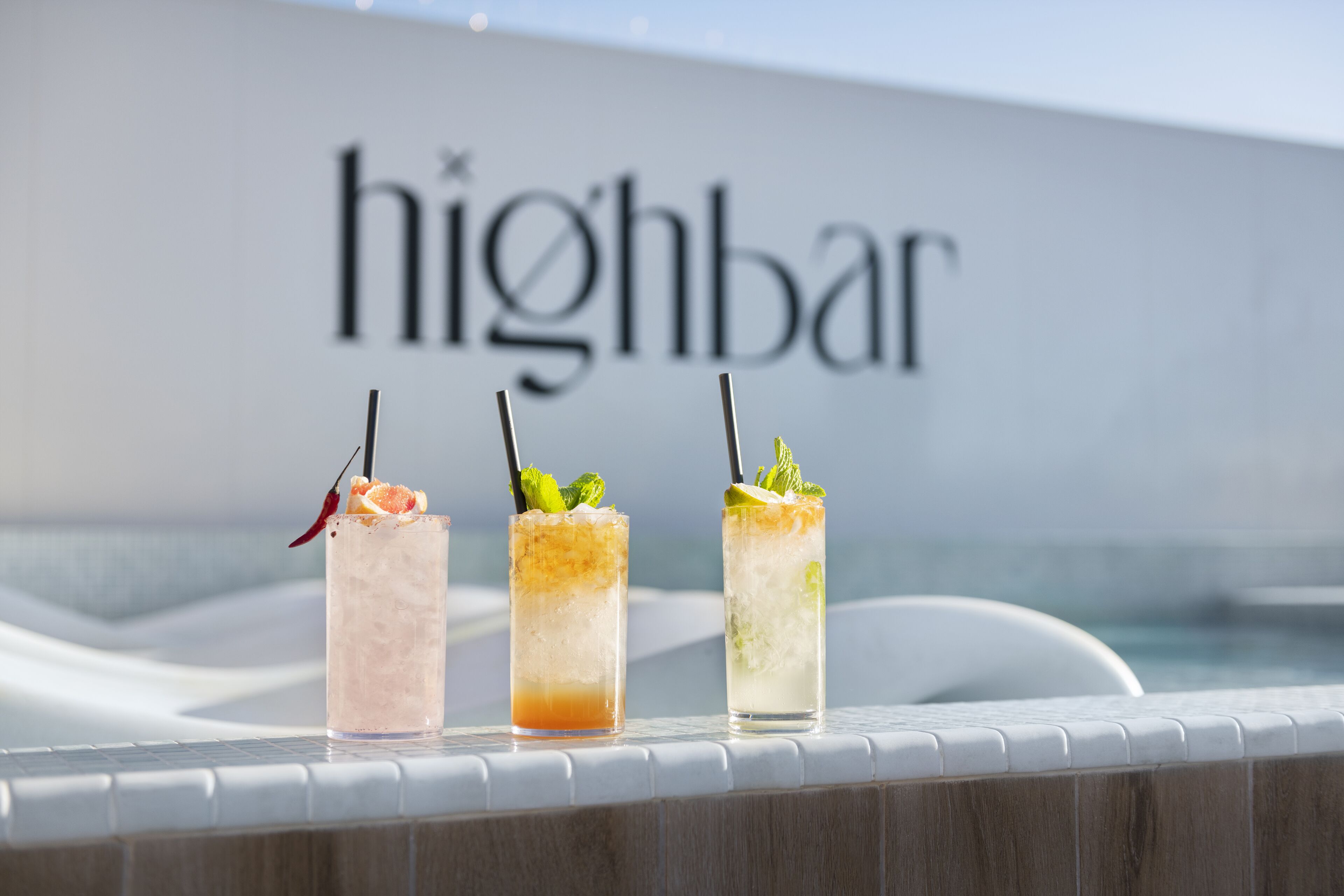 Tres cócteles sobre una barra junto a la piscina, con un letrero que dice 'highbar' al fondo.