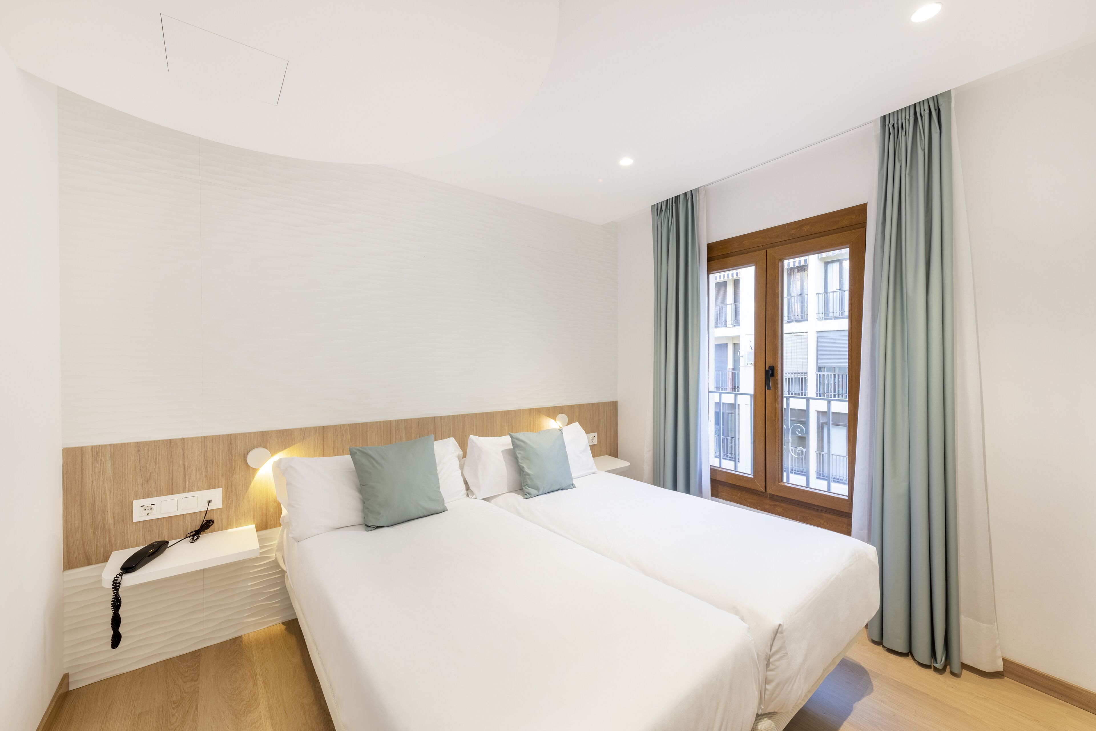 A hotel room with two beds, a wooden headboard, a bedside telephone, and a window with curtains.
