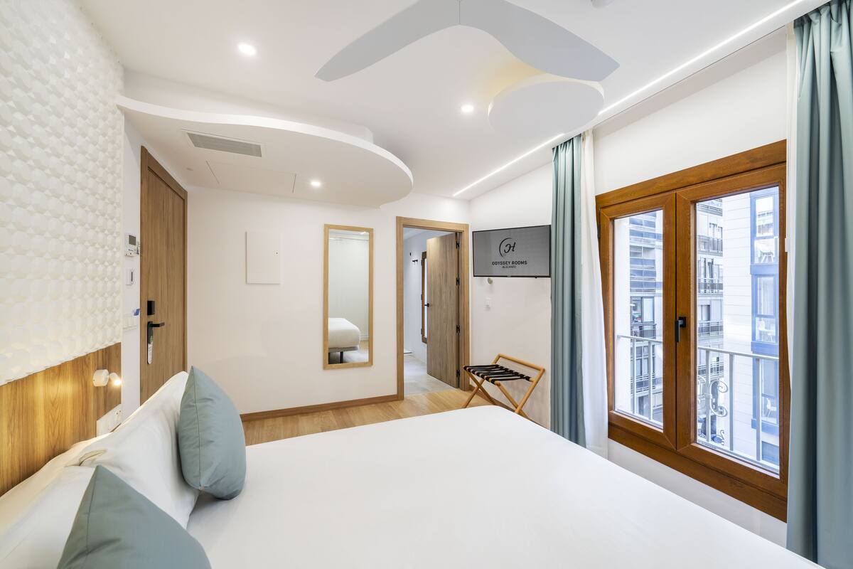 Ein modernes Hotelzimmer mit einem großen Bett, einem Holzkopfstück, einem Spiegel, einer Holztür und Blick auf die Stadt durch ein Fenster.