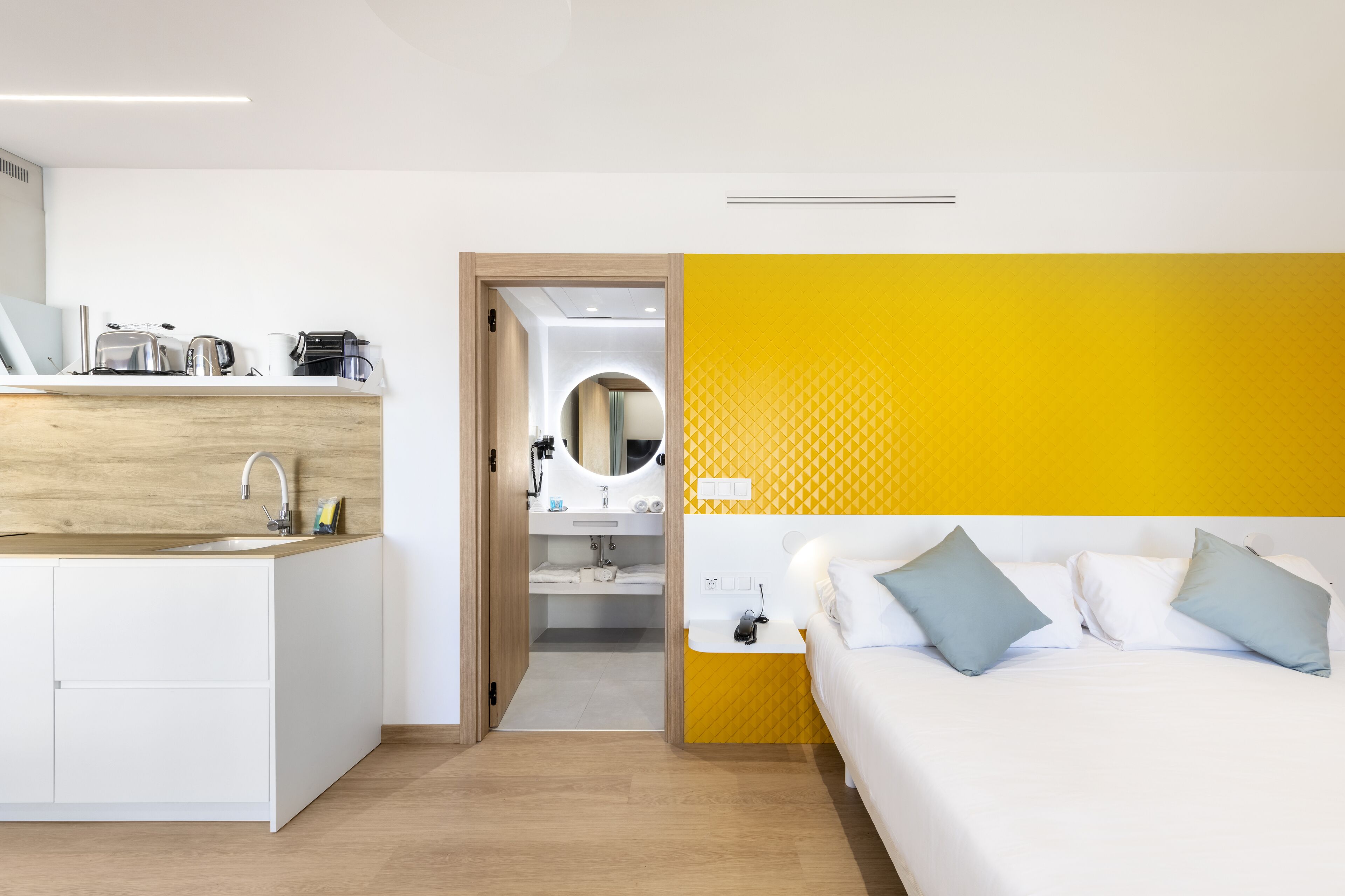 A modern hotel room with a kitchen area, a bed with pillows, and a bathroom visible through an open door.