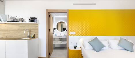 A modern hotel room with a kitchen area, a bed with pillows, and a bathroom visible through an open door.