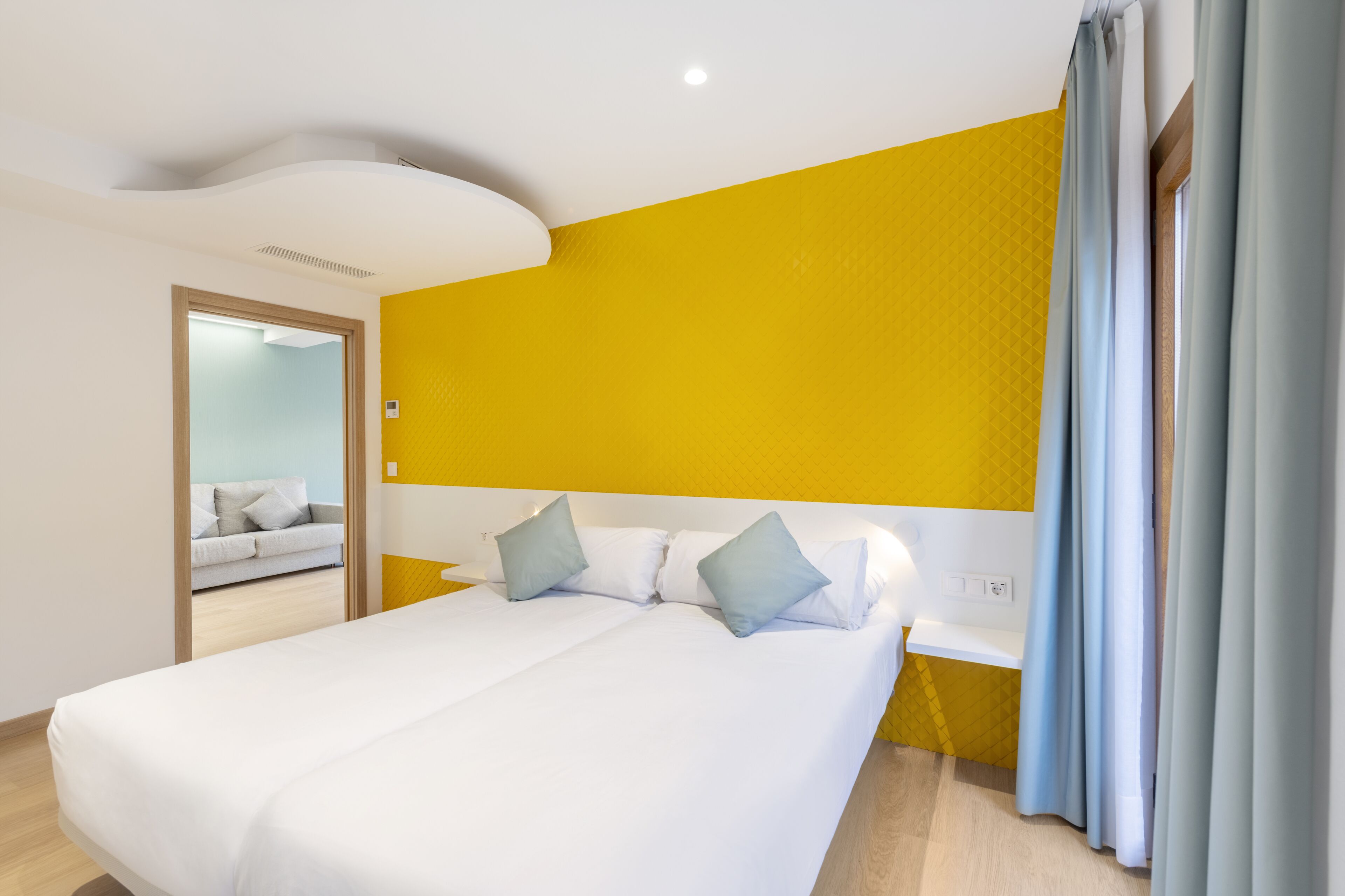 Ein modernes Hotelzimmer mit einem großen Bett, einem Nachttisch und Blick auf einen Flur mit einer Couch.