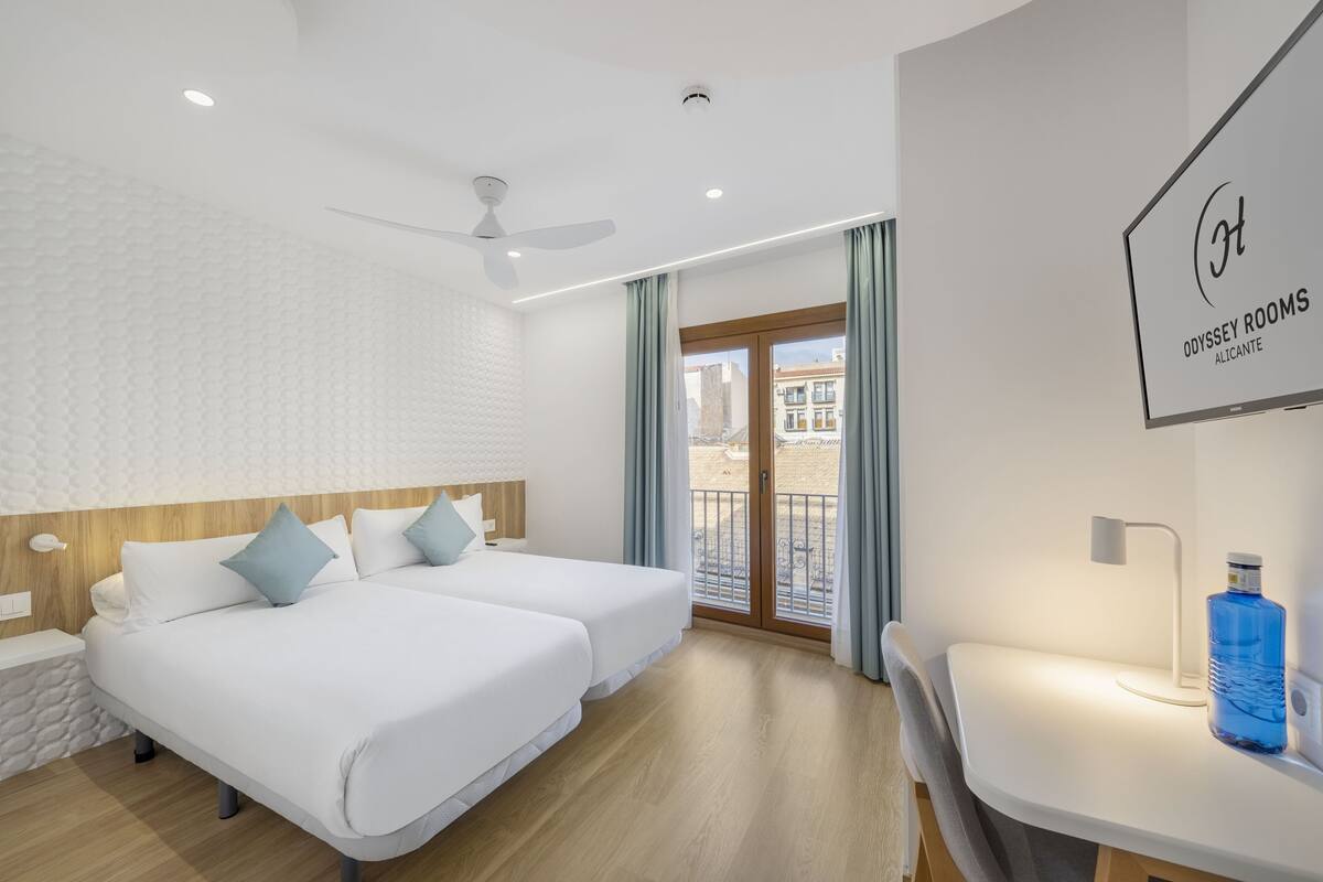 Ein Hotelzimmer mit zwei Betten, einem Schreibtisch, einem Stuhl, einem Deckenventilator und Blick auf die Stadt vom Balkon.