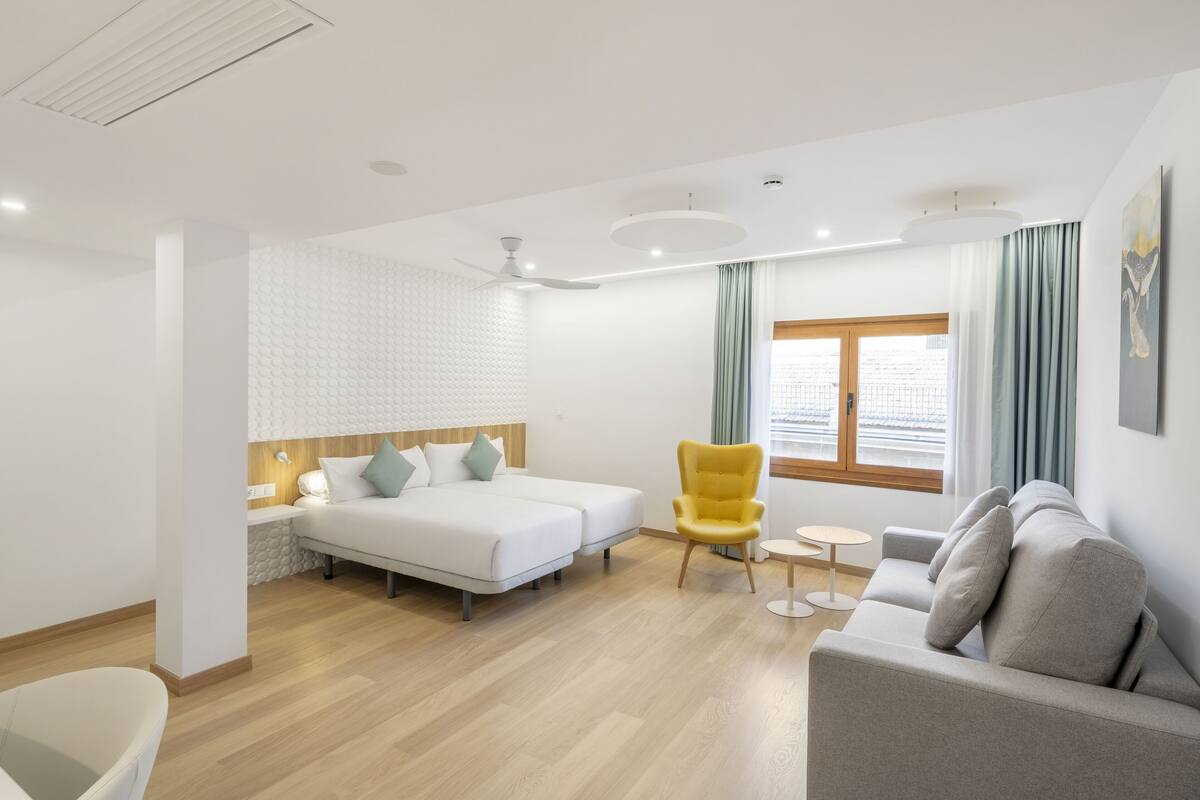 Ein modernes Hotelzimmer mit Bett, einer Couch, einem gelben Sessel, einem kleinen runden Tisch und einem Fenster mit Vorhängen.
