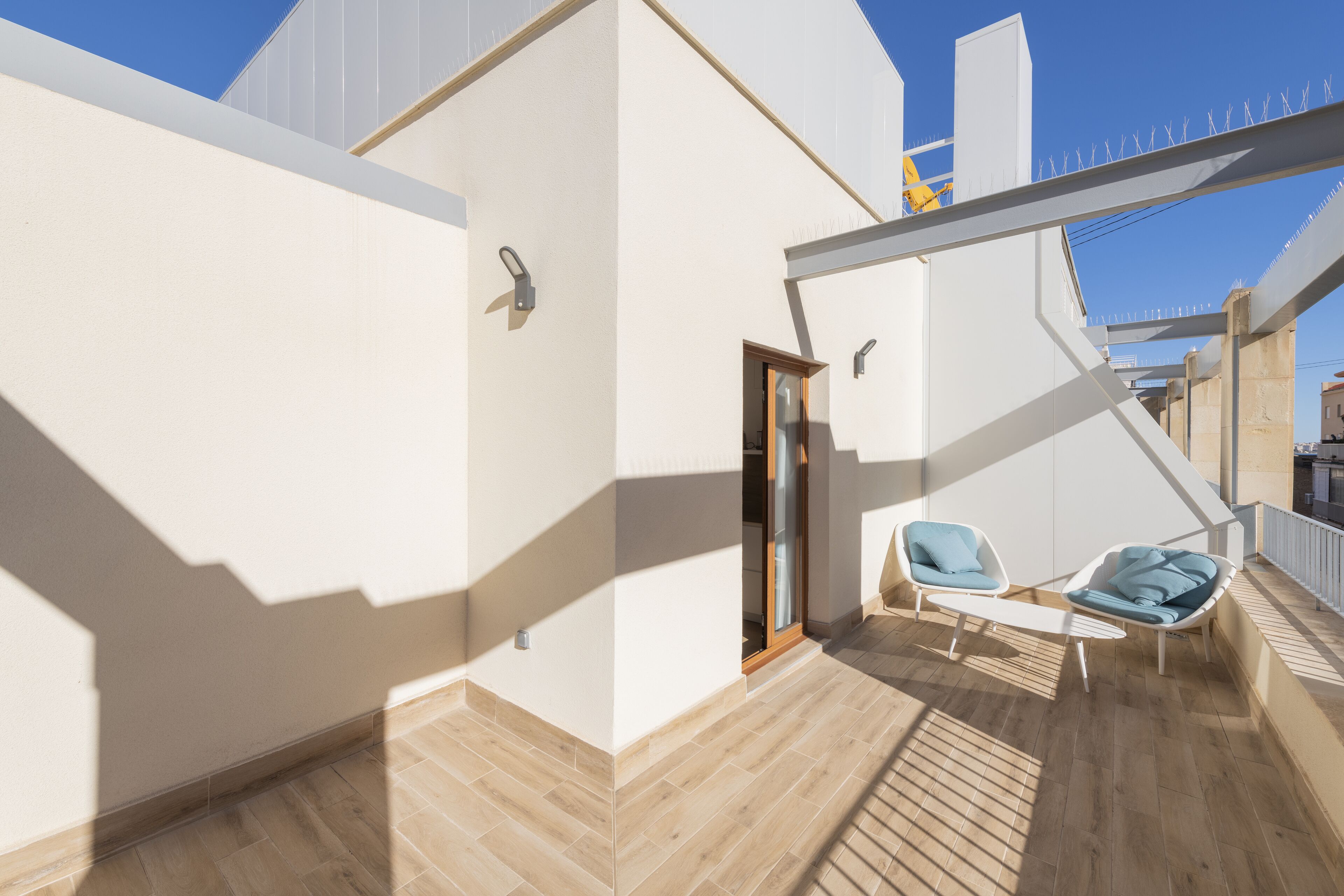Un balcón moderno al aire libre con piso de madera, dos sillones azules y una puerta que da a un espacio interior.