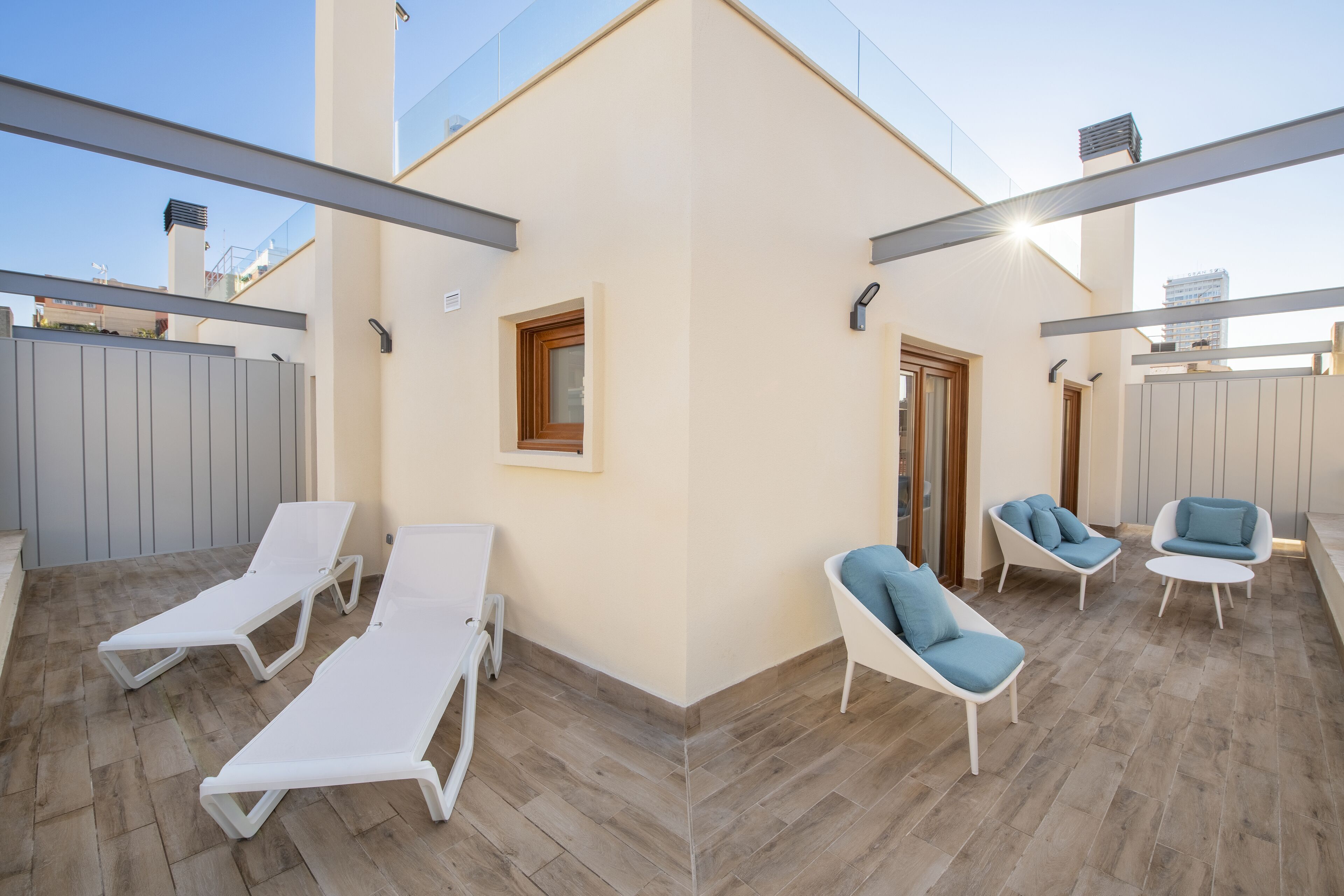 Un patio exterior con sillones blancos y sillas con cojines azules, una puerta de madera y vistas del horizonte de la ciudad.