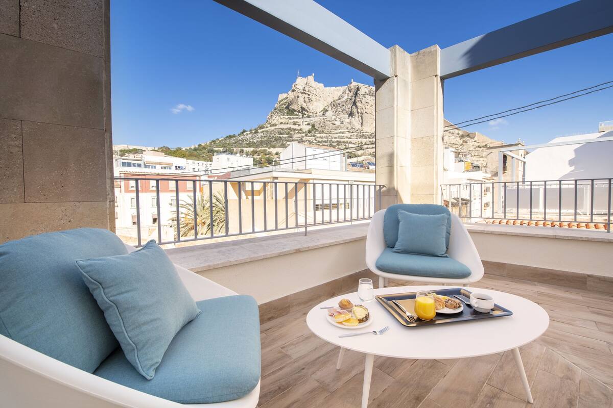 Ein Balkon mit einer Couch und einem Sessel, ein Tisch mit Frühstücksgebäck und Blick auf einen Berg.