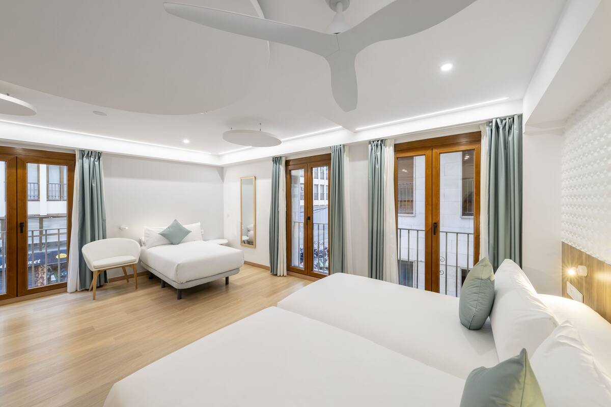 Ein modernes Hotelzimmer mit einem großen Bett, einer Sitzecke mit Stuhl und kleinem Tisch sowie einem Balkon mit Vorhängen.