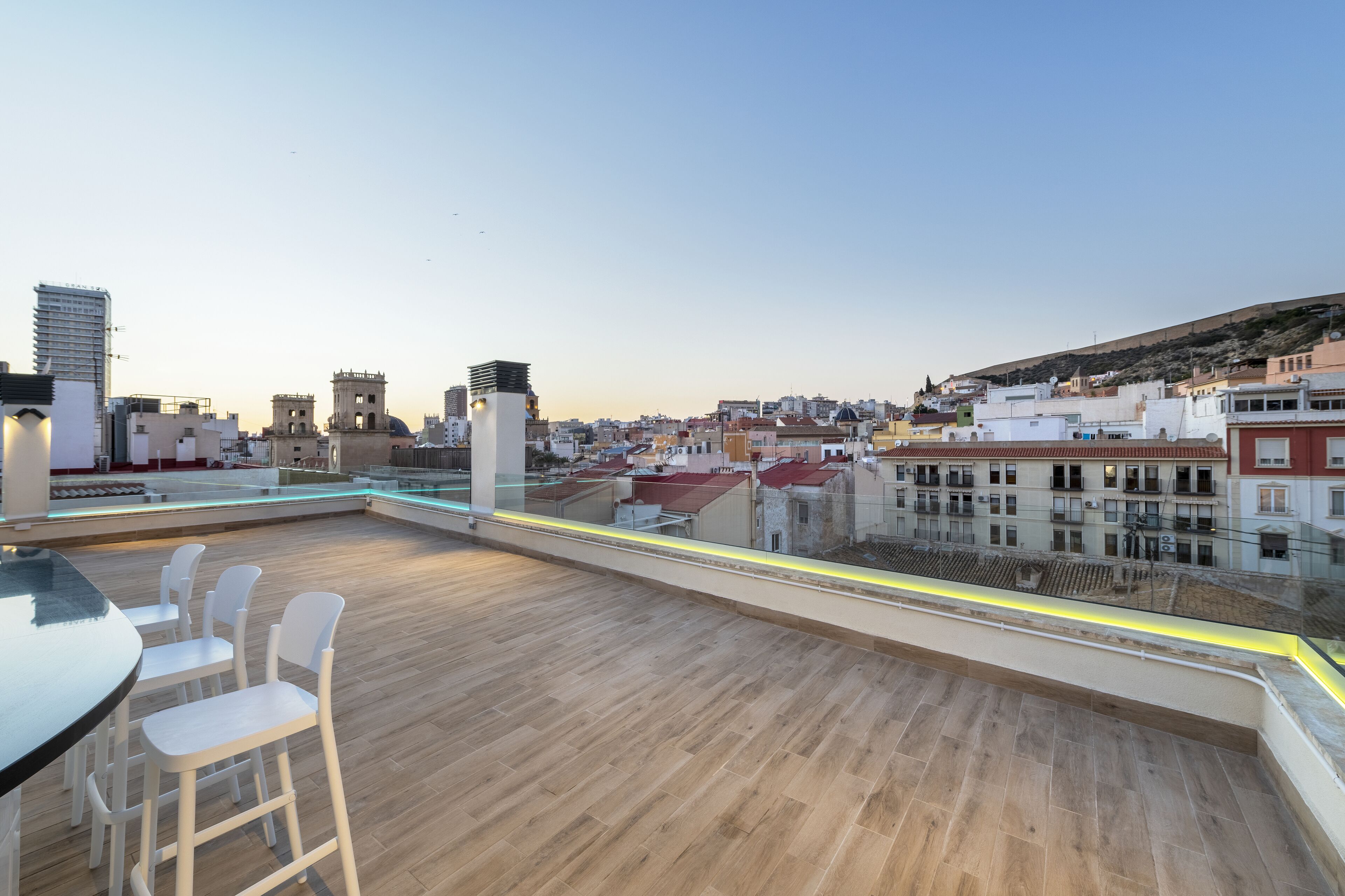 Terraza en la azotea con una mesa y sillas, que ofrece vistas panorámicas de la ciudad y una colina al fondo.