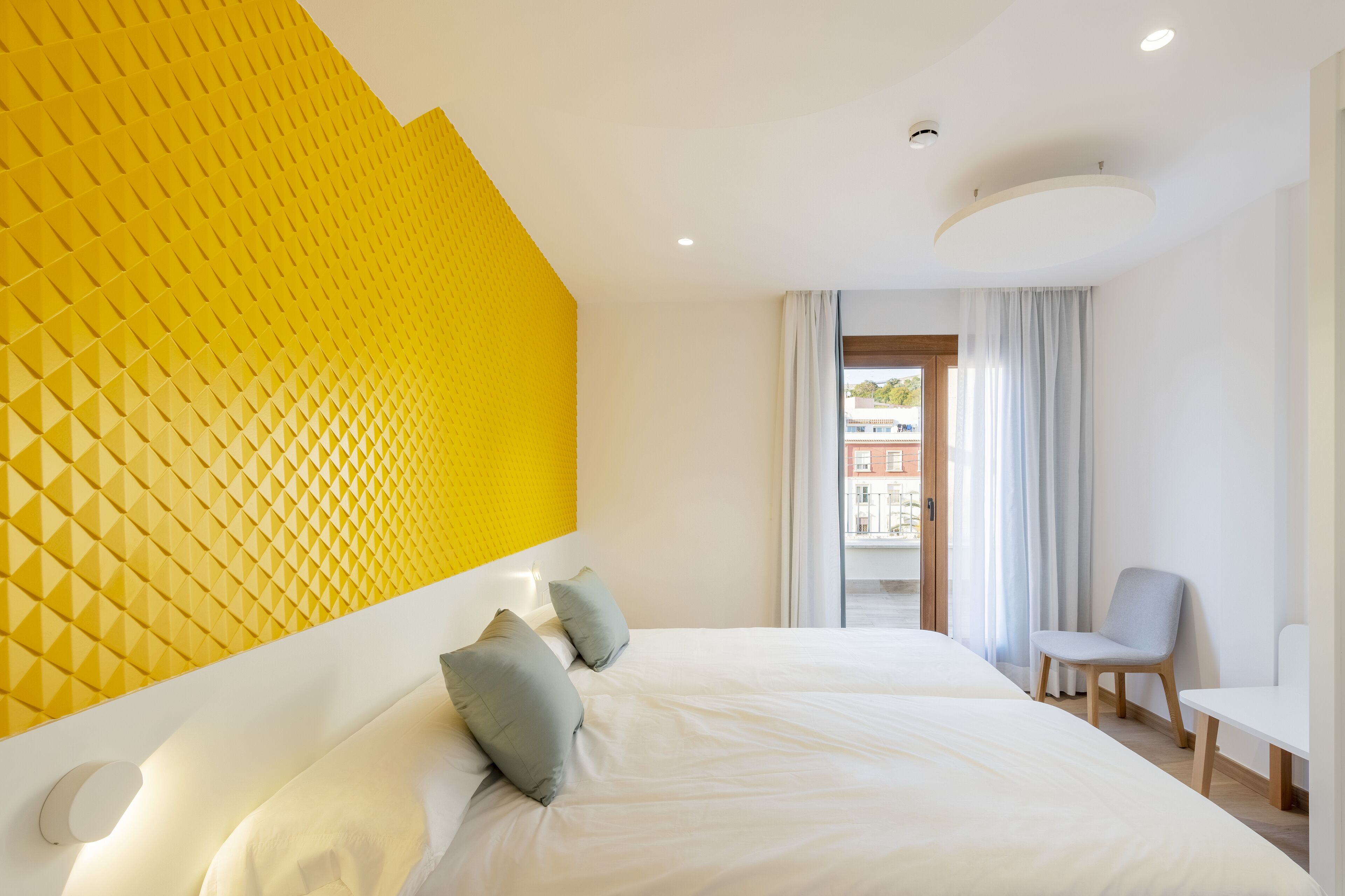 A hotel room with a large bed, a bright yellow accent wall, a small seating area, and a view of the outside.
