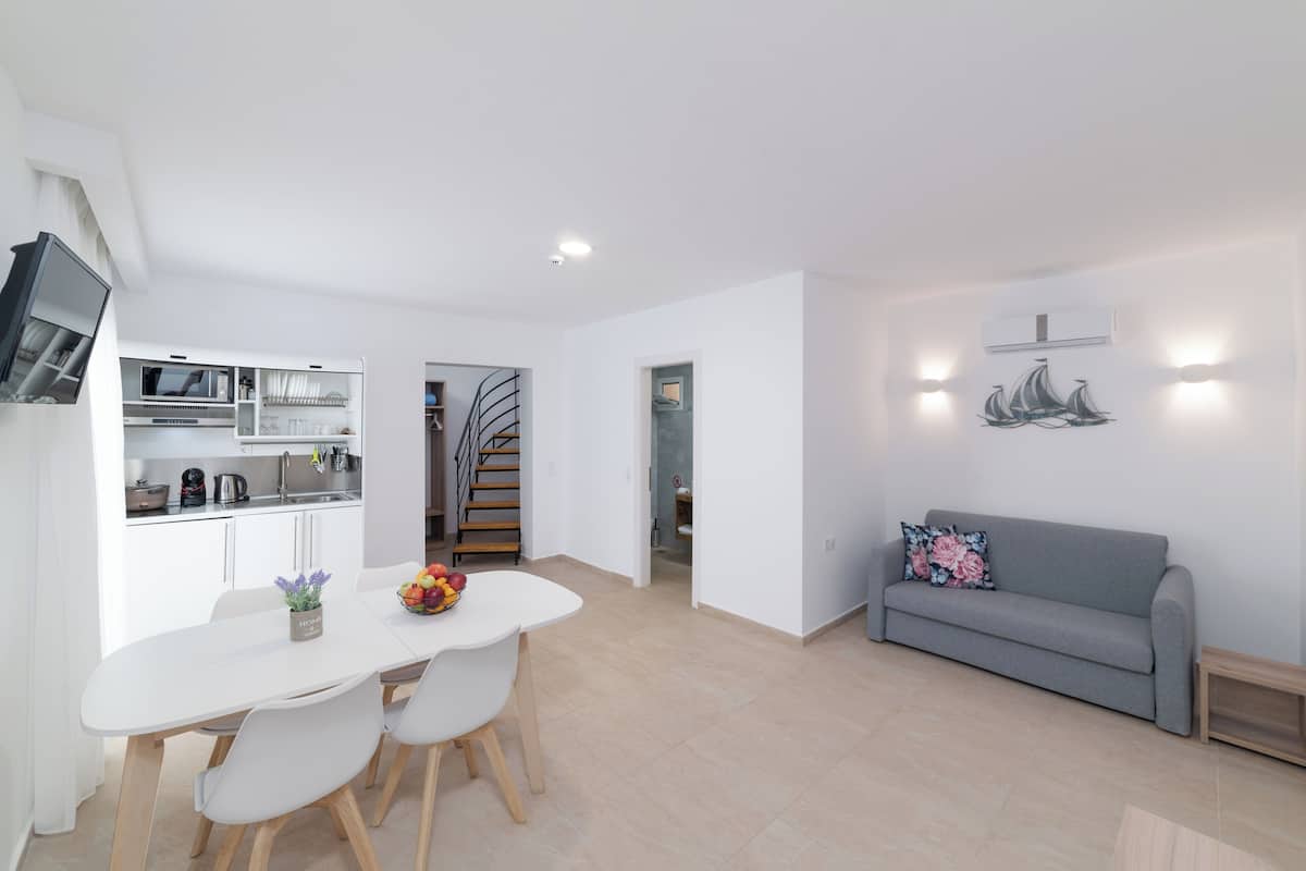 Una cocina moderna con armarios blancos, una mesa de comedor con sillas y un sofá gris en una sala de estar luminosa con paredes blancas y una escalera.