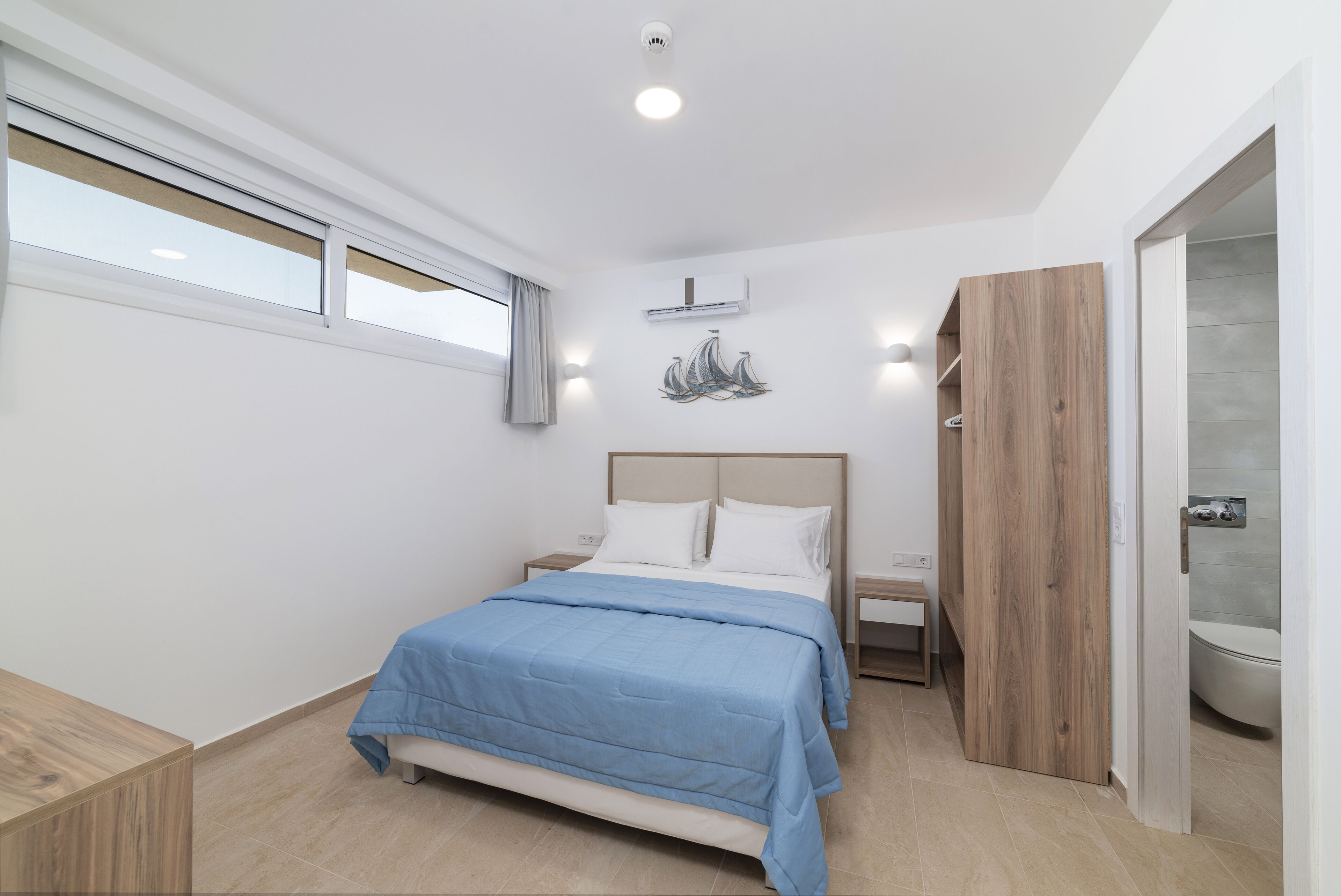 Un dormitorio moderno con una cama, mesitas de noche, un armario de madera y un baño visible a través de una puerta abierta.