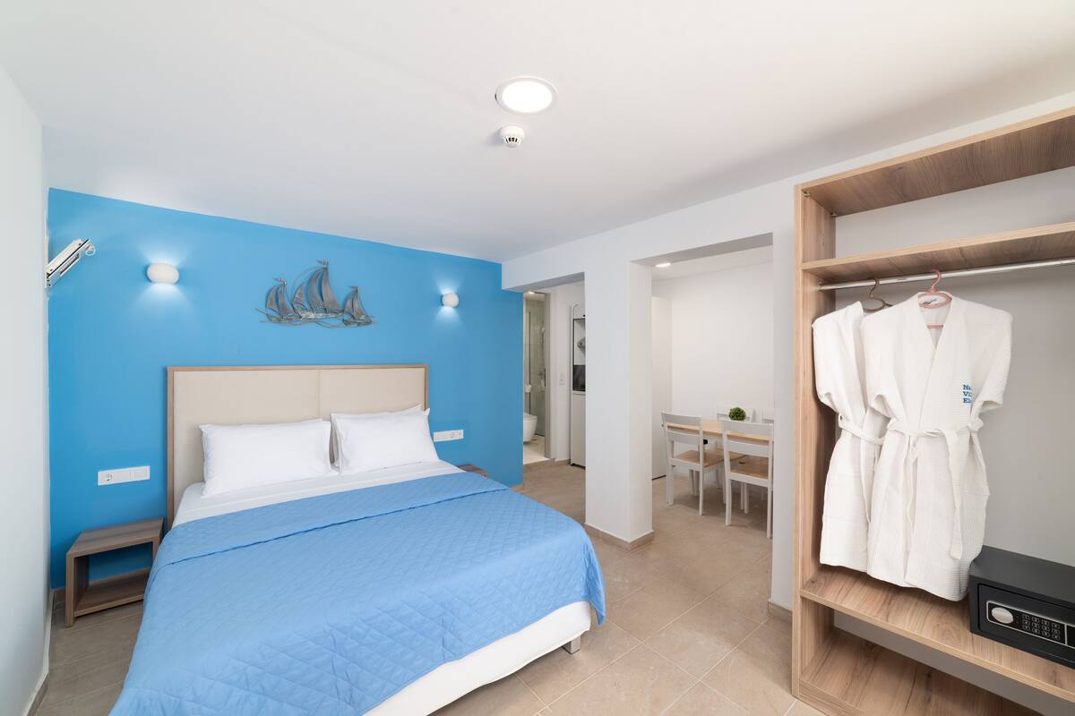 Habitación de hotel moderna con una pared azul de acento, una cama con colcha azul, una mesita de noche de madera, una bata colgada en un perchero de madera y una pequeña mesa con una silla.