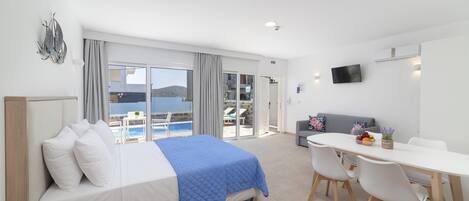 A modern hotel room with a bed, a dining table with chairs, a TV, and a view of the sea.