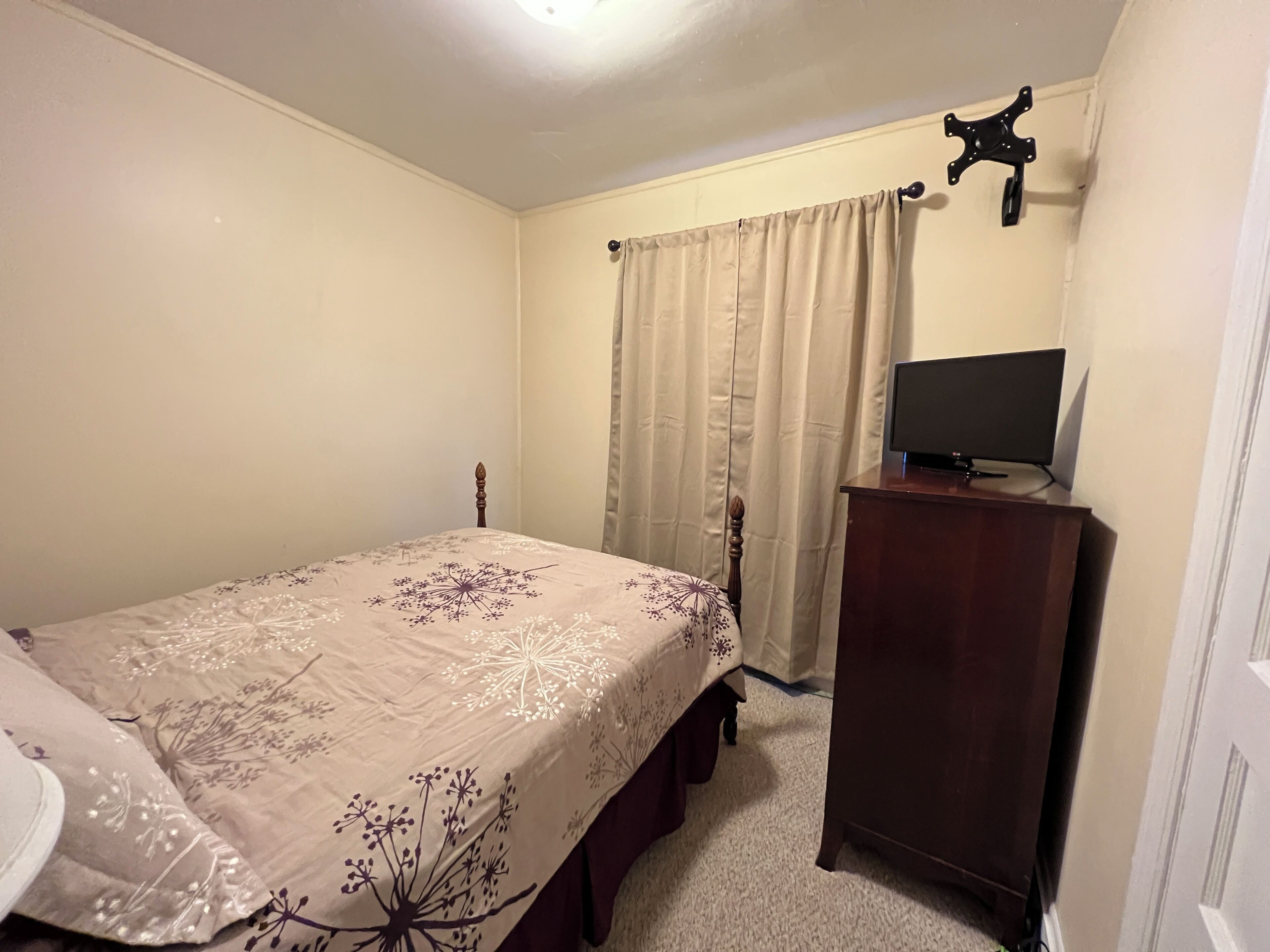 A bedroom with a bed, a wooden dresser, a television on the dresser, and a window with curtains.