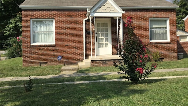 A two-story brick house with a front porch, a garage door, and a small garden with pink flowers. - Harlan, Ky Modern Private Guesthouse 304 Historic Downtown (Vela's Villas) (Harlan)