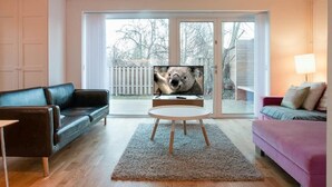 Een woonkamer met een zwarte leren bank, een roze bank, een ronde salontafel en een televisie op een standaard.
