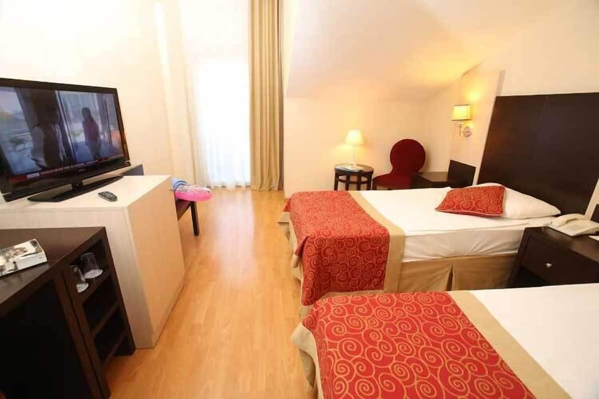 A hotel room with a TV, two beds with red and white bedding, a desk with a lamp, and a chair.