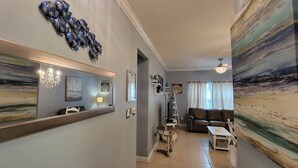 A spacious living room with a large sofa, a coffee table, and a chandelier. There is a staircase leading to an upper level, and a large painting on the wall.