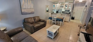 A living room with a brown leather sofa, a small white table, and a dining area with a blue tablecloth. - EZ Walk to Beach * Swanky 2 bedroom * Sleeps Tons (Mar y Sol II #1) by RedAwning (South Padre Island)