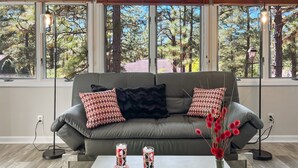 Una sala de estar moderna con un sofá gris, cojines estampados, una mesa de centro con bebidas y un control remoto, y vistas a árboles a través de amplios ventanales.