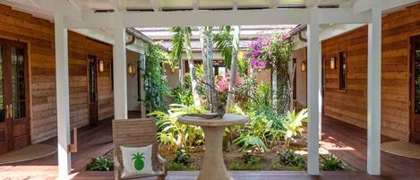 A wooden interior with a central round table and a chair, surrounded by lush tropical plants and a wooden ceiling structure.