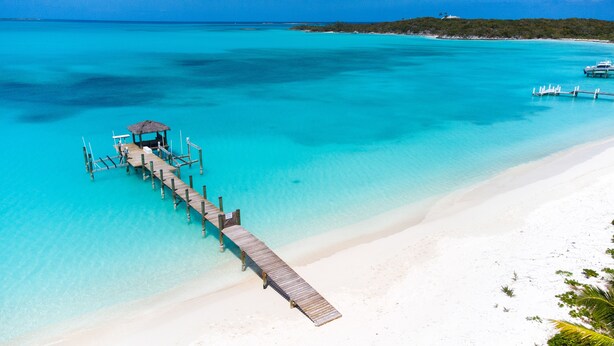 Une jetée en bois mène à une structure à toit de chaume, située sur une plage de sable blanc où l’eau est d’un bleu turquoise cristallin.